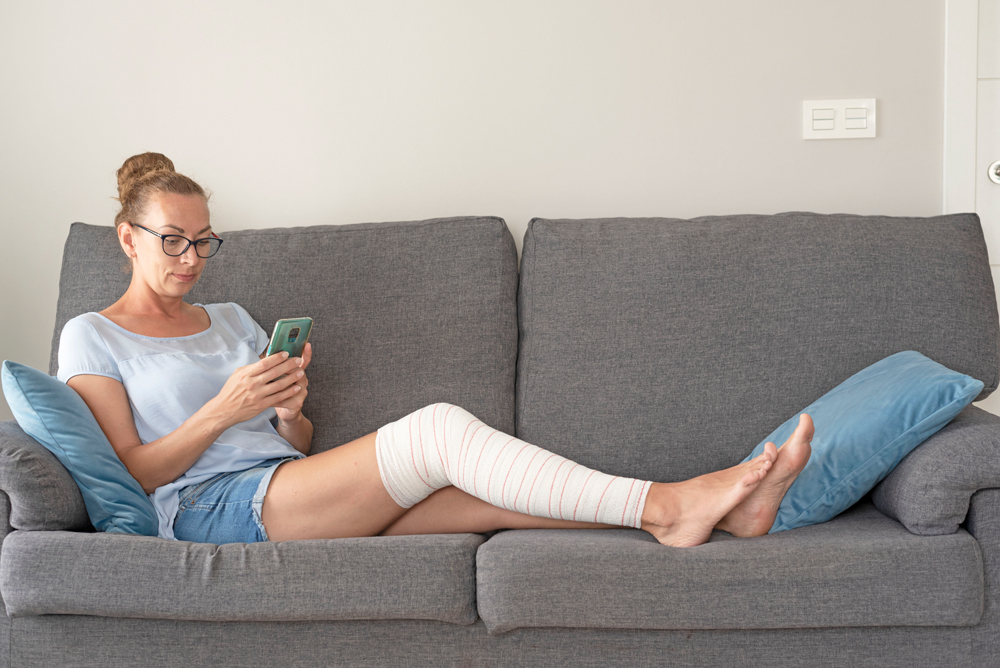 young-woman-sitting-sofa-with-broken-leg-bandage-looking-mobile-phone